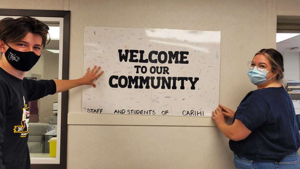 Two high school students stand in front of a poster they made that says "welcome to our community". The poster contains 300 signatures from fellow students.