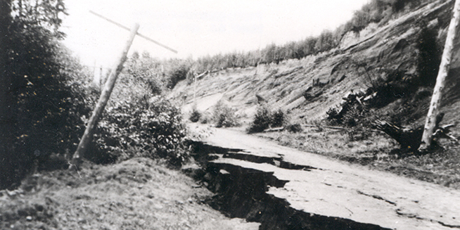 An old, black and white photograph of a road that has been broken by an earthquake. A phone pole leans over on the left.