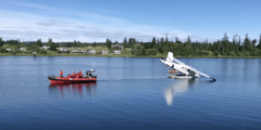 A damaged white floatplane is towed across the water by a red dingy.