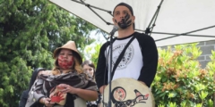 Ken Watts speaks outside in front of a microphone. He is holding a drum and has a black handprint across his mouth. He is standing next to Cynthia Dick, who wears a woven cedar hat.