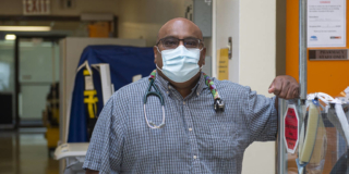 Dr. Prean Armogam leans on some medical equipment. He is wearing a mask and has a stethoscope hung around his neck.