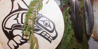 A Huu-ay-aht drum hangs next to cedar branches and feathers.