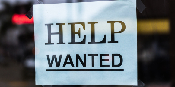 A "Help wanted" sign is taped inside a restaurant window.