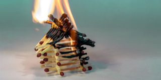 A toy house built out of matches burns in front of a pale green background.