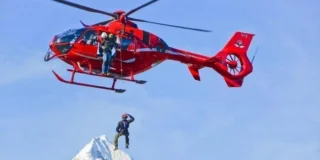 A red helicopter lowers someone onto a mountain on a sunny day.