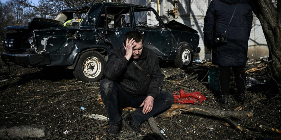 A man crouches with his head in his hands in front of his bombed-out car.