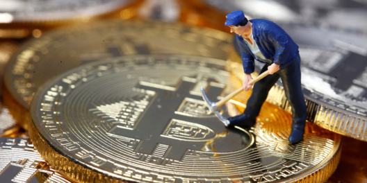 A tiny figure of an old-fashioned gold digger stands on top of a gold Bitcoin.