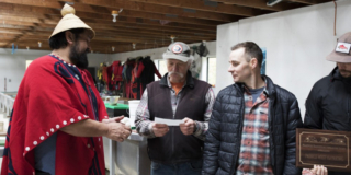 Tyee Ha'wilth Hasheukumiss, wearing a traditional woven hat and bright red cape, hands a $10,000 cheque to Doug Paulfrey, Tofino Salmon Enhancement Society manager.