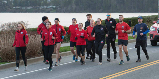 A group of runners in red shirts and black bottoms run along a road next to a lake. The weather is not very good.