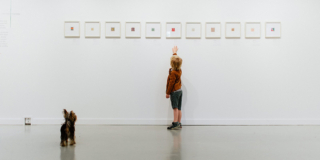 A small child reaches up to touch a painting that is hung on a white wall. There is a tiny dog at his feet.