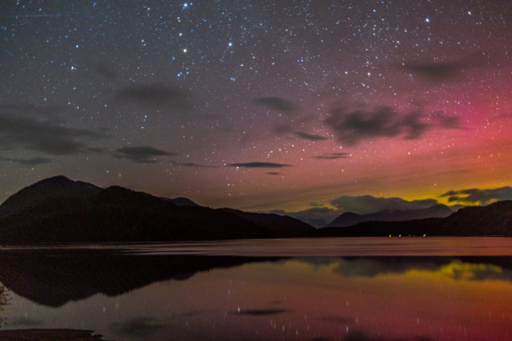 A night picture of pink and yellow northern lights over low mountains in Tofino. There are bright stars in the sky.