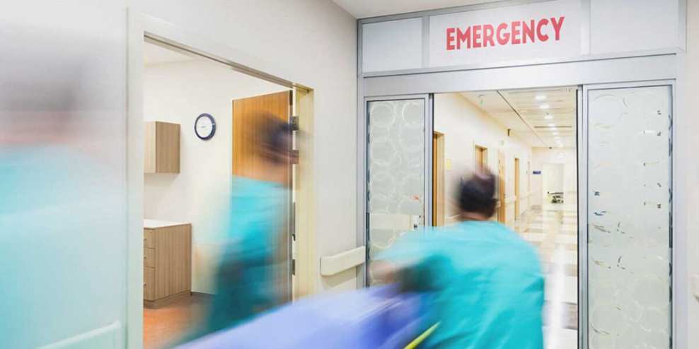 Healthcare workers rush a patient on a gurney toward a door that reads EMERGENCY. The picture is blurry.