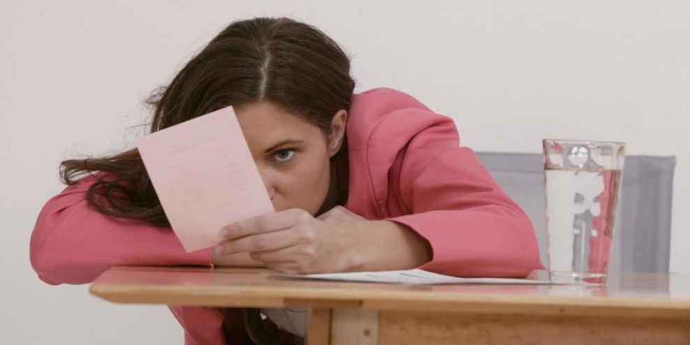 A woman leans over an office table and looks depressed. Part of her face is obscured by a pink slip.