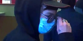 A closeup of a man with a hood, mask, and sunglasses taken from a security camera.