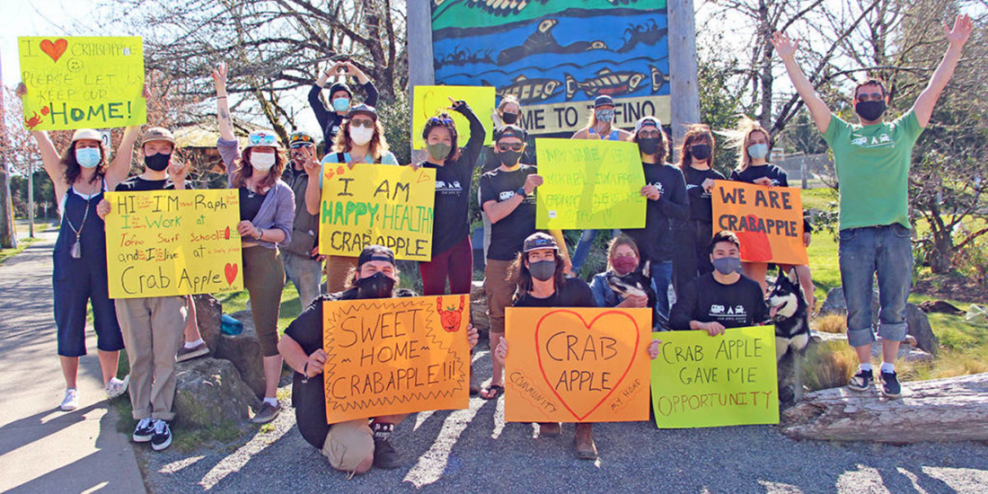 A group of Crab Apple Campground residents stand in front of the Tofino welcome sign with posters that proclaim how the campground has helped them.