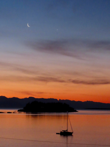 A crescent moon hangs over a sailboat at sunrise.