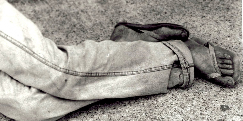 A black-and-white photo of a person lying on a sidewalk. The photo is cropped at the person's knees. Their jeans and feet are dirty.