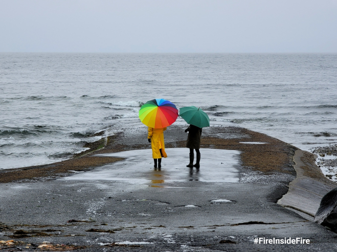 Two people with bright umbrellas stand on a grey and windy beach in Courtenay, BC.
