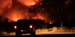 An SUV sits with its lights on in the dark against a wildfire burning in the background.