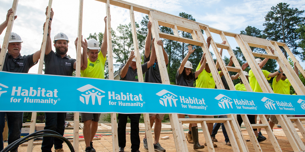 A group of people in hard hats raise a new wall with a sign on it for Habitat for Humanity.