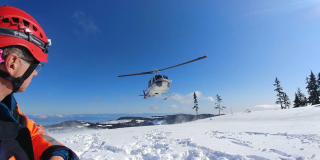 A member of the search and rescue stands on a snowy mountain facing a helicopter that is taking off.