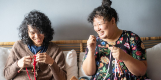 Two women sit on a couch together and laugh and knit.