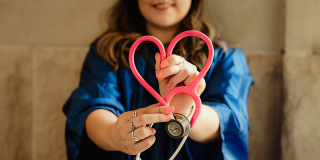 A woman in a graduation robe holds a stethoscope in front of her that's been bent to resemble a heart.