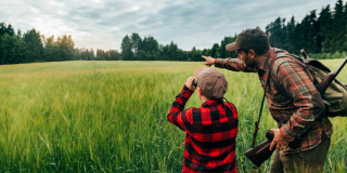 A man stands holding a hunting rifle in a green field with his son who is looking through binoculars.