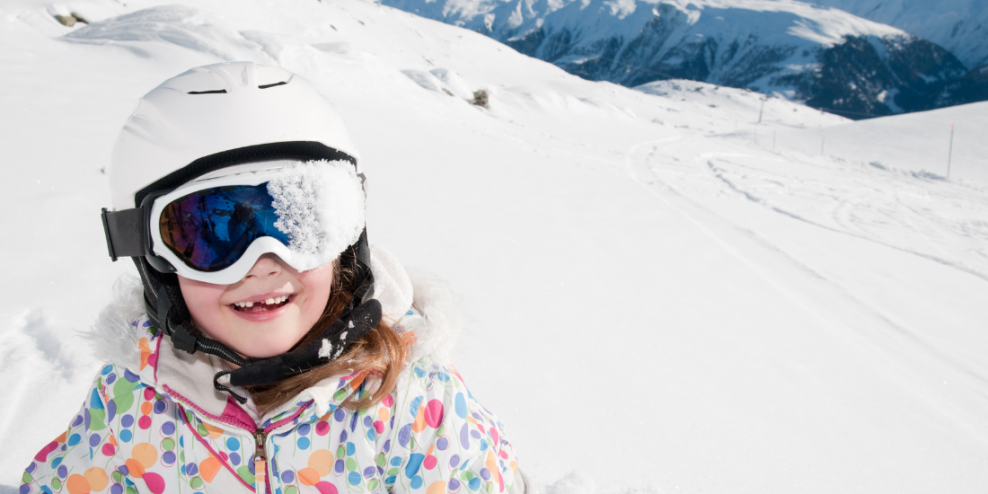 A young skier smiles for the camera on a snowy mountain. One side of her goggles is covered in snow.