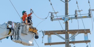 Two technicians in a cherry picker inspect equipment at the top of a hydro pole on a sunny day.