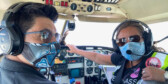 Teara Fraser and her co-pilot sit in the cockpit of a small airplane. They're wearing cloth masks with Indigenous designs and Fraser is wearing aviator sunglasses.