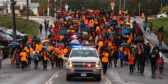 A large group of people in orange shirts march along a street behind a police vehicle in the rain.