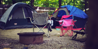 Two foldable camping chairs sit in front of tents in a green, shady campsite.