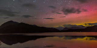 A night picture of pink and yellow northern lights over low mountains in Tofino. There are bright stars in the sky.