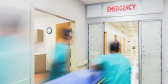 Healthcare workers rush a patient on a gurney toward a door that reads EMERGENCY. The picture is blurry.