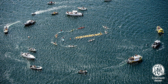 A drone shot of boats circling on the water around a floating sign that reads "Fish Farms Out."