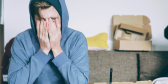 A young guy in a blue hoody sits on a couch with his head in his hands. There are empty boxes around him.