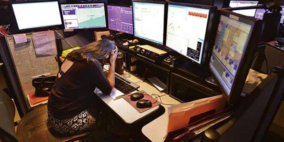 A tired 911 dispatcher sits with her head in her hands in front of six screens.