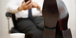 A business man scrolls through his phone with his feet on the table. The photo is cropped to cut out his face and his feet are closest to the camera.