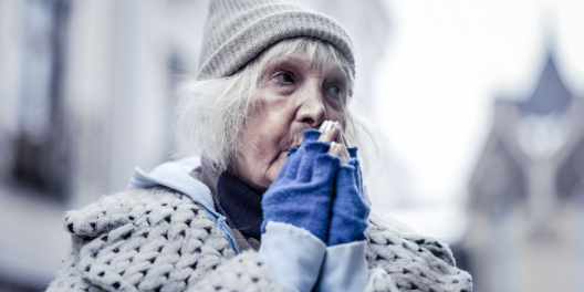 An older woman in a toque and gloves blows into her hands to warm them up.