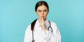 A healthcare worker in a white coat makes a "shhhh" finger over their mouth and looks at the camera with concern.