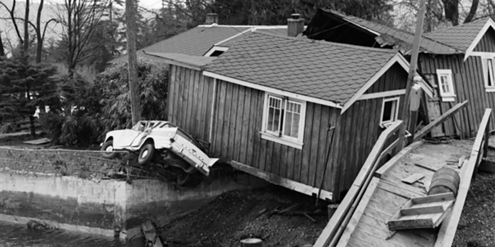 A black-and-white photo of a house that's been knocked off its foundations with a car underneath it.