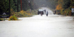 People stand on the other side of a flooded section of road in front of the Pathfinder RV Park sign in the rain.