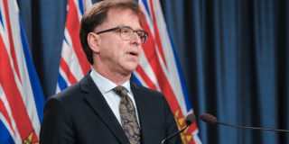 Adrian Dix stands in front of a podium with BC flags in the background. He looks tired.