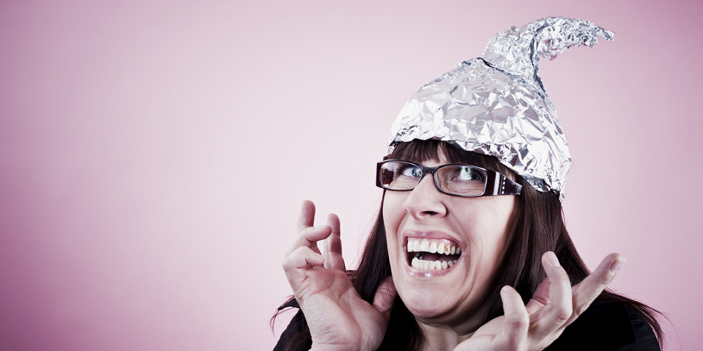 A woman looks nervous in a tinfoil hat against a shiny pink background.