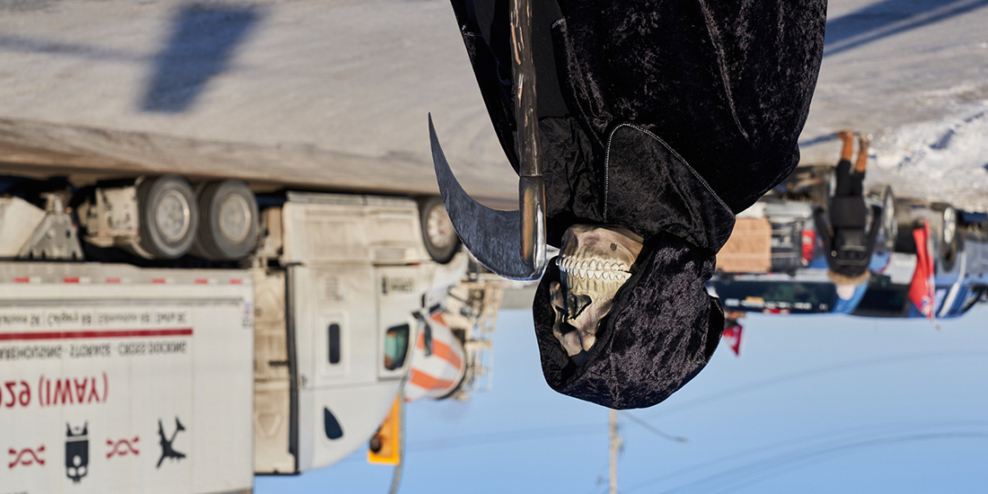 A person stands dressed as the Grim Reaper in front of a truck on a rural highway.