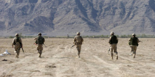 Five soldiers walk away from the camera toward mountains in a desert.