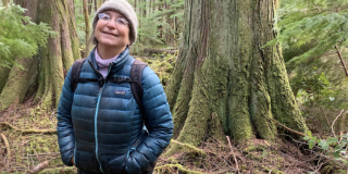 A woman in a blue puffy jacket and knit toque smiles in front of a huge mossy tree.