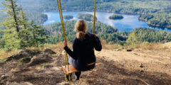A wowan sits on a swing tied to a tree at the top of a forested hill. The view looks out over a lake.