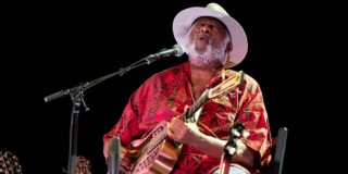 Legendary blues musician sings and plays his guitar on stage. He's wearing a bright red shirt and a white cowboy hat.